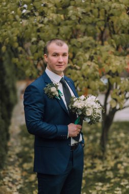 Mavi takım elbiseli bir adam elinde bir buket çiçek tutuyor. Gülümsüyor ve kameraya bakıyor.