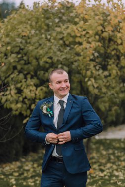 Mavi takım elbiseli ve kravatlı bir adam gülümsüyor ve kravatını düzeltiyor. Yerde yaprakları olan bir ağacın önünde duruyor.