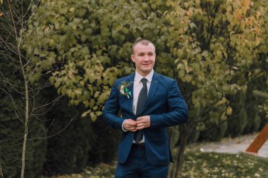 Mavi takım elbiseli ve kravatlı bir adam bir ağacın önünde duruyor. Yakasında bir çiçek ve parmağında bir yüzük var. Formalite ve zarafet kavramı