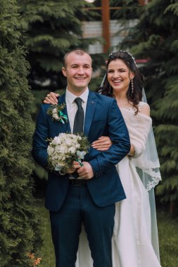 Bir adam ve bir kadın ormanda fotoğraf için poz veriyorlar. Adam mavi bir takım elbise ve kravat takıyor ve kadın beyaz bir elbise giyiyor. İkisi de gülümsüyor ve mutlu görünüyorlar.