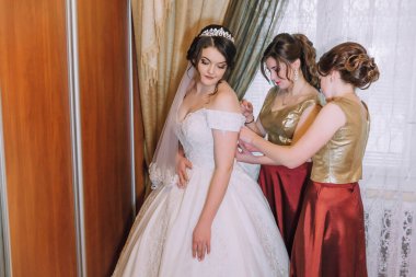 Üç kadın düğün için hazırlanıyor. İçlerinden biri beyaz bir elbise ve taç giyiyor. Diğer ikisi kırmızı elbiseler giyiyor. Tahta bir kapının önünde duruyorlar.