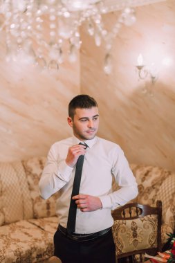 Bir adam kravatını bağlamaya hazırlanıyor. Bir koltuğun ve sandalyenin önünde duruyor.