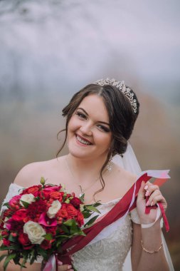 Bir kadın taç takıyor ve elinde bir buket kırmızı çiçek tutuyor. Gülümsüyor ve mutlu.