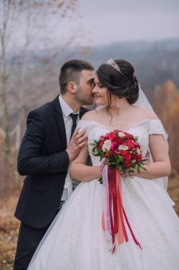 Bir gelin ve damat bir buket kırmızı çiçeğin önünde birbirlerinin dudaklarını öpüyor.