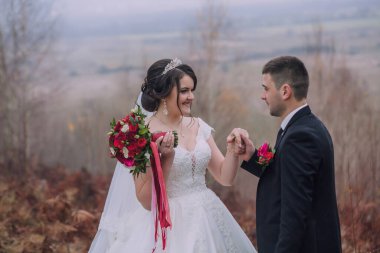 Bir gelin ve damat ormanda el ele tutuşup gülümsüyor. Damat takım elbise giyerken gelin beyaz bir elbise ve kırmızı bir buket giyiyor. Sahne romantik ve resimli.