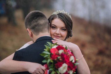 Bir gelin ve damat bir buket kırmızı çiçek tutarken birbirlerine sarılıyorlar. Gelin taç takıyor ve damat da takım elbise giyiyor. Sahne samimi ve romantik, aşkı yakalıyor.