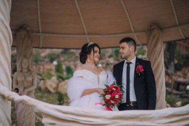 Gelin ve damat çadırın altında, gelin bir buket tutuyor. Sahne romantik ve mutlu.