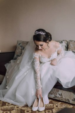 Bir gelin kanepede oturuyor, düğün ayakkabılarını giyiyor. Gelin büyük günü için hazırlanırken imajın romantik ve samimi bir havası var.