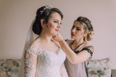 Bir kadın saçını başka bir kadına yaptırıyor. Gelin beyaz bir elbise ve taç giyiyor.