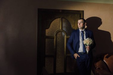 Takım elbise giymiş ve kravat takmış bir adam elinde bir buket çiçekle kapının önünde duruyor. Birini bekliyor ya da ayrılmaya hazırlanıyor. Elindeki çiçekler pembe ve beyaz karışımı.