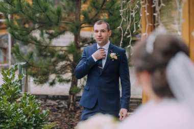 Mavi takım elbiseli bir adam kravatını bağlamaya hazırlanıyor. Arkasında bir kadın duruyor, izliyor.