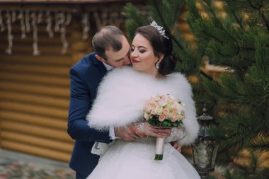Bir gelin ve damat ormanda birbirlerine sarılıyorlar. Gelin bir kürk manto giyiyor ve elinde bir buket çiçek tutuyor.