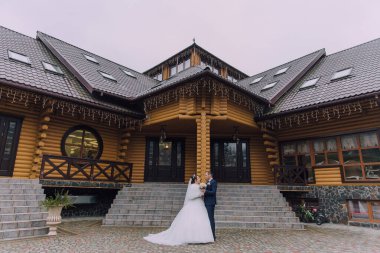 Bir gelin ve damat büyük bir ahşap evin önünde duruyor. Gelin beyaz bir elbise giyiyor ve damat mavi bir takım elbise giyiyor. İkisi de gülümsüyor ve mutlu görünüyorlar. Evin bir köylüsü var.