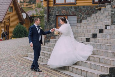 Gelin ve damat merdivenlerde duruyorlar, gelin beyaz elbise giyiyor ve damat takım elbise giyiyor.