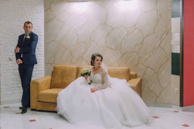 Bir gelin ve damat kanepenin önünde fotoğraf için poz veriyorlar. Gelin beyaz bir elbise giyiyor ve elinde bir buket tutuyor. Adam takım elbise ve kravat takıyor. Sahne mutlu ve romantik.