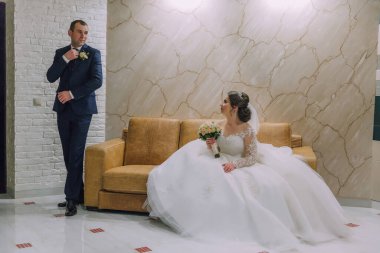 Bir adam ve bir kadın kanepede oturuyorlar. Adam takım elbise ve kravat takıyor. Kadın gelinlik giyiyor.