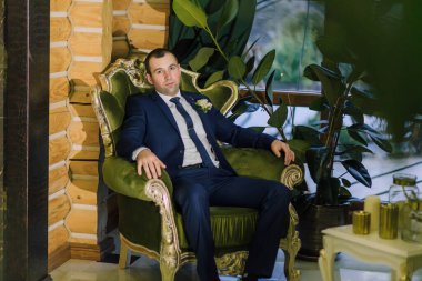 Takım elbiseli bir adam yeşil bir sandalyede oturuyor. O bir kravat ve yaka çiçeği takıyor.