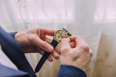 Bir adam saat takıyor ve ayarlıyor. Saat altın ve siyah. Adam mavi bir takım elbise giyiyor.