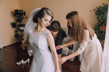 Bir gelin, iki kadının yardımıyla düğününe hazırlanıyor. Sahne bir kutlama ve destektir.