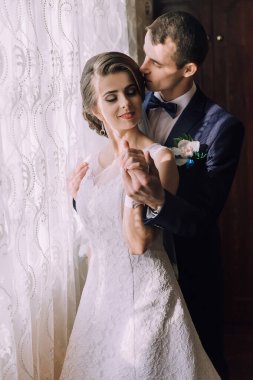 Bir gelin ve damat pencerede birbirlerine sarılıyorlar. Gelin beyaz bir elbise giyiyor ve damat mavi bir takım giyiyor.