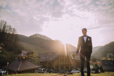 Takım elbiseli bir adam elinde bir buket çiçekle evin önünde duruyor. Sahne, arka planda güzel bir dağ manzaralı kırsal bir alanda geçiyor.