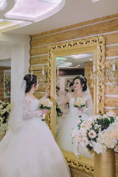 Gelinlik giymiş bir kadın aynanın önünde elinde bir buket çiçek tutuyor. Ayna süslenmiş ve altın renginde ve oda çiçeklerle süslenmiş. Zarafet ve kutlama kavramı