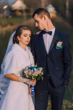 A bride and groom are posing for a picture. The bride is wearing a white dress and a white shawl, while the groom is wearing a black suit. They are holding a bouquet and a bouquet holder, respectively clipart