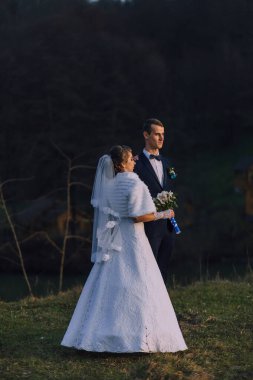 Gelin ve damat çimenli bir yamaçta dururlar, gelin beyaz bir elbise giyer ve damat mavi bir takım giyer.