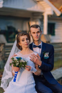 Bir gelin ve damat bir evin önünde fotoğraf için poz veriyorlar. Gelin bir buket tutuyor ve damat kravat takıyor. Sahne mutlu ve romantik.