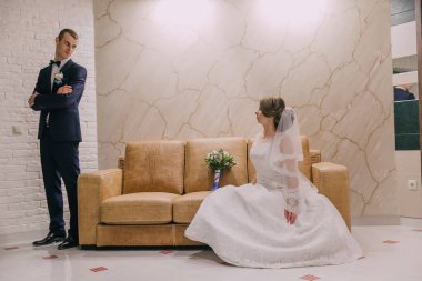 Bir adam ve bir kadın kanepede oturuyorlar. Adam takım elbise giyiyor ve kadın da gelinlik giyiyor. Adam suratında kaşları çatık olan kadına bakıyor. Sahne gergin ve garip.