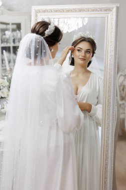 Bir kadın düğünü için hazırlanıyor ve aynada kendine bakıyor. Beyaz bir elbise ve taç giyiyor.