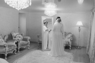 Bir gelin büyük aynalı bir odada düğününe hazırlanıyor. Bir sandalyenin ve lambanın önünde duruyor.