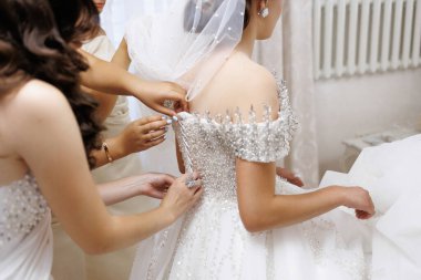 Üç kadın düğün için hazırlanıyor. Bir tanesi gelinin elbisesini ayarlıyor. Gelin üzerinde altın kolye olan beyaz bir elbise giyiyor. Diğer iki kadın hazırlanmasına yardım ediyor.