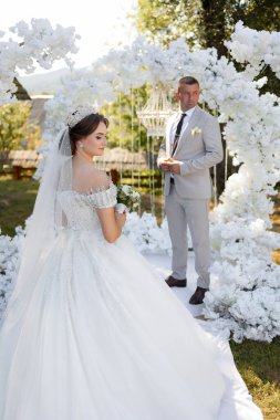 Bir gelin ve damat beyaz bir kemerin önünde duruyor. Gelin, taç ve saçında çiçek olan beyaz bir elbise giyiyor. Damat takım elbise ve kravat takıyor. Sahne bir düğün töreni.