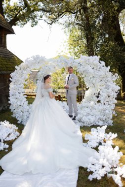 Bir gelin ve damat beyaz bir kemerin önünde duruyor. Gelin beyaz bir elbise giyiyor ve damat bir takım elbise giyiyor. Kemer yolu beyaz çiçeklerle süslenmiş.