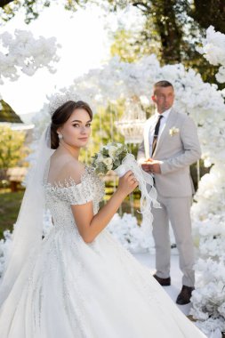 Bir gelin ve damat beyaz bir kemerin önünde duruyor. Damat takım elbise giyerken gelin bir taç ve beyaz bir elbise giyiyor. Gelin bir buket tutuyor.
