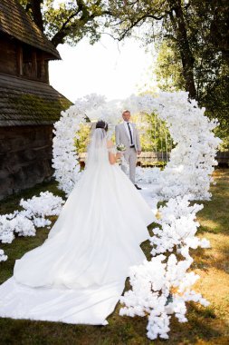 Bir gelin ve damat beyaz bir kemerin önünde duruyor. Gelin beyaz bir elbise giyiyor ve damat bir takım elbise giyiyor. Kemer yolu beyaz çiçeklerle süslenir, romantik bir hava yaratır.