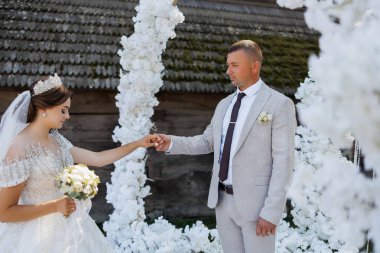 Bir gelin ve damat beyaz bir kemerin önünde duruyor. Gelin bir buket tutuyor ve damat kravat takıyor. Sahne muhtemelen bir düğün merasimi.