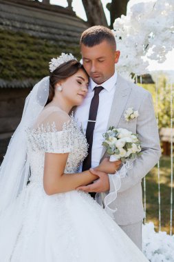 Bir gelin ve damat beyaz bir kemerin önünde poz veriyorlar. Gelin taç takıyor ve damat kravat takıyor. Sahne mutlu ve romantik.