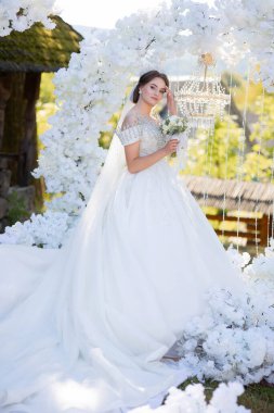 Gelin, peçeli beyaz bir elbise içindeki bir resim için poz veriyor. O bir buket çiçek tutuyor. Sahne arka planda beyaz kemerli bir bahçede geçiyor.