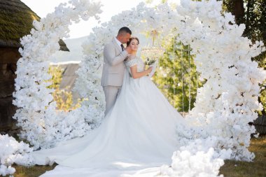 Bir gelin ve damat beyaz bir kemerin önünde duruyor. Gelin bir buket tutuyor ve damat onu yanağından öpüyor. Sahne bir düğün töreni.