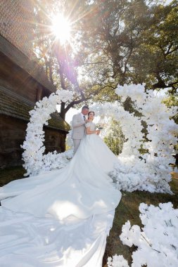 Bir gelin ve damat beyaz bir kemer yolunun önünde poz veriyorlar. Gelin uzun beyaz bir elbise giyiyor ve damat bir takım elbise giyiyor. Kemer yolu beyaz çiçeklerle süslenmiş.