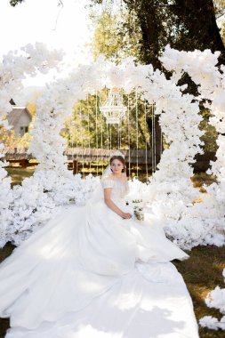 Beyaz bir kemer yolunun önünde beyaz elbiseli bir kadın oturuyor. Kemer yolu beyaz çiçeklerle süslenmiş ve üzerinde bir avize var. Kadın beyaz bir gelinlik giyiyor.