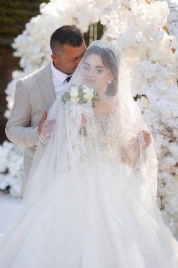 Bir gelin ve damat beyaz bir kemerin önünde poz veriyorlar. Gelin peçe takıyor ve damat onu öpüyor. Gelin elinde bir buket çiçek tutuyor.