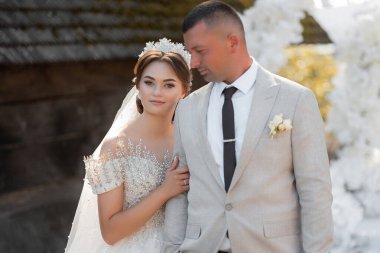 Bir gelin ve damat binanın önünde fotoğraf için poz veriyorlar. Gelin beyaz bir elbise ve taç giyerken damat takım elbise ve kravat takıyor. Sahne mutlu ve kutlama dolu.