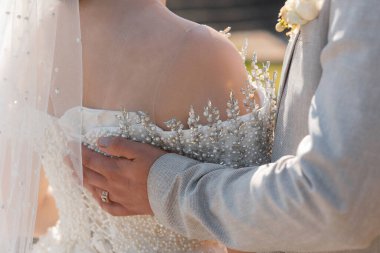 Gelin ve damat birbirlerini kucaklıyor, gelin beyaz elbise giyiyor ve damat gri takım elbise giyiyor. Gelinin elbisesi inci ve tacla süslenmiş.