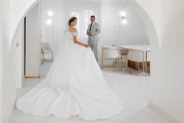 Beyaz elbiseli bir gelin takım elbiseli bir adamın önünde duruyor. Oda beyaz mobilyalarla süslenmiş ve modern bir tasarıma sahip.