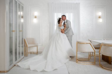 Bir gelin ve damat altın koltuklu beyaz bir odanın önünde öpüşüyorlar. Oda beyaz mobilyalarla süslenmiş ve modern bir tasarıma sahip.