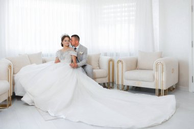 Bir gelin ve damat bir odada kanepede oturuyor. Gelin uzun beyaz bir elbise giyiyor ve damat bir takım elbise giyiyor. Oda beyaz mobilyalarla süslenmiş ve temiz, zarif bir görünüme sahip.