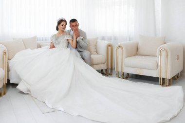 Bir gelin ve damat, arka planda büyük beyaz bir kanepe olan bir odada, bir kanepede oturuyor.